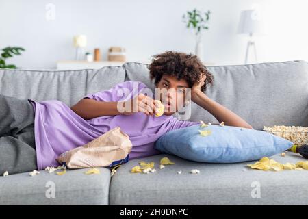 Adolescent afro ennuyé allongé sur un canapé avec des en-cas dispersés, manger des frites, regarder un spectacle terne ou un film à la télévision à la maison Banque D'Images