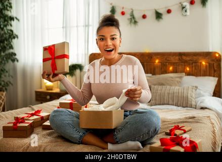 Portrait de la dame noire recevant des cadeaux de Noël et déballant la boîte en carton, assis sur le lit dans la chambre confortable Banque D'Images