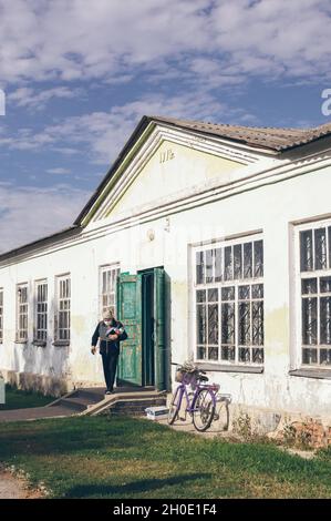 Une femme masquée sort d'un vieux bâtiment d'époque qui s'écaille de 1971 par une journée ensoleillée, un vélo contre le mur Banque D'Images