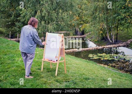 Une femme heureuse tire sur le chevalet avec une brosse et des peintures. Femme artiste dessine la nature et les arbres sur papier au bord de l'eau sur la rive de la rivière Banque D'Images