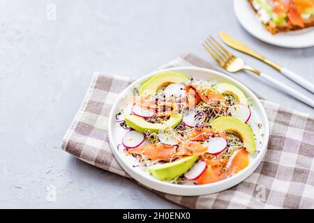 Flat Laissez sur le dessus une salade saine avec du saumon fumé, de l'avocat, du radis et des pousses de luzerne fraîches sur une assiette blanche sur du béton gris Banque D'Images