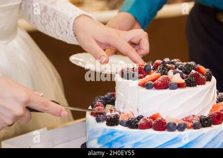 La mariée et le marié ont coupé un gâteau blanc et bleu crème à deux niveaux lors d'une fête de mariage.Gâteau décoré de baies sauvages. Banque D'Images