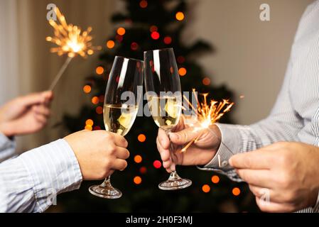 Couple de verres à champagne et de tenir des spamerons sur le fond de l'arbre de noël, célébrant Noël à la maison, gros plan. Banque D'Images