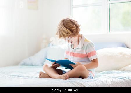 Les enfants lisent un livre dans le lit.Chambre pour enfant.Un petit garçon lit des livres sur un lit blanc avec une literie colorée.Enfants en pépinière.Enfant garçon jouant à l'intérieur. Banque D'Images