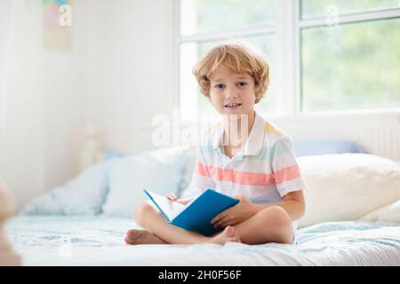 Les enfants lisent un livre dans le lit.Chambre pour enfant.Un petit garçon lit des livres sur un lit blanc avec une literie colorée.Enfants en pépinière.Enfant garçon jouant à l'intérieur. Banque D'Images