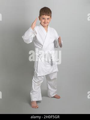 Garçon dans un kimono blanc sourit et se tient en position défensive de combat sur fond gris. Banque D'Images