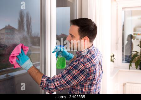 Beau jeune homme nettoyant les fenêtres avec un chiffon et une bouteille vaporisatrice à la maison Banque D'Images