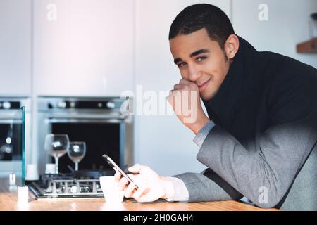 Un jeune homme amusé se souriant derrière sa main en s'inclinant sur une table de cuisine tenant son téléphone portable et tournant pour regarder l'appareil photo Banque D'Images