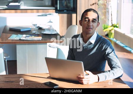 Jeune Noir regardant avec un sourire la caméra pendant qu'il est assis à une table de travail sur un ordinateur portable dans un puits de lumière du soleil Banque D'Images