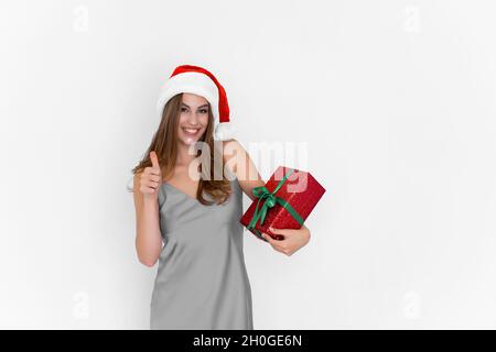 Joyeux sourire positif Santa fille tient boîte cadeau tandis que les gestes pouce vers le haut faisant excellent.Célébrez. Banque D'Images