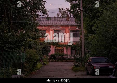 Nuit photo de la petite ville ancienne maison de deux étages cour noyée dans la verdure avec une vieille voiture garée sur le côté de la route au crépuscule avec la lune au-dessus des nuages Banque D'Images