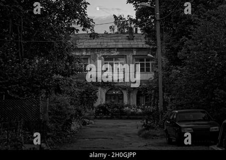 Photo en plein air noir et blanc d'une petite ville ancienne maison de deux étages cour noyée dans la verdure avec une vieille voiture garée sur le côté de la route au crépuscule avec la lune Banque D'Images