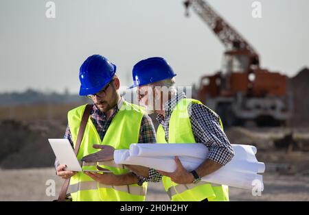 Deux ingénieurs regardent un ordinateur portable sur le chantier avec des machines de construction en arrière-plan Banque D'Images