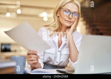 Femme d'affaires mûre et concentrée discutant de documents pendant une visioconférence en ligne à l'aide d'un ordinateur portable tout en étant assise à son bureau Banque D'Images