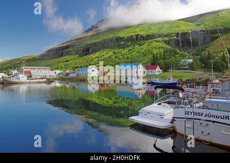 Bateaux et petites maisons reflétés dans le fjord, cascade sur le flanc de montagne vert, Seydisfjoerdur, Islande Banque D'Images