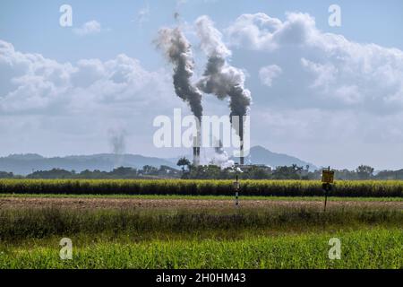 Un moulin à canne à sucre en saison de broyage avec des cheminées émettant de la fumée dans le ciel nuageux et une récolte de canne à sucre au premier plan Banque D'Images