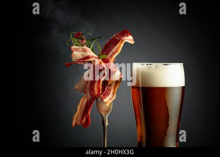 Un verre de bière et des tranches de bacon frit savoureux garni de romarin.Arrière-plan noir avec espace de copie. Banque D'Images