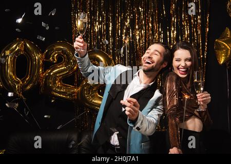 couple élégant et gai avec des verres à champagne et des sparkers près de la décoration de noël dorée sur le noir Banque D'Images