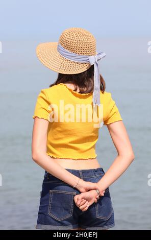 jeune fille skinny avec chapeau de paille et mains derrière elle debout au bord de la mer Banque D'Images