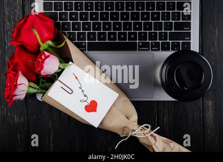 vue du dessus un bouquet de roses rouges en papier artisanal avec carte postale attachée sur un ordinateur portable avec une tasse de papier de café sur fond de bois sombre Banque D'Images