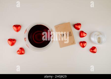 vue de dessus d'un verre de vin avec des bonbons au chocolat en forme de coeur enveloppés de papier d'aluminium rouge et une petite carte postale sur fond blanc Banque D'Images