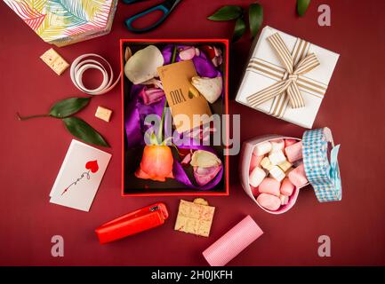 vue de dessus d'une boîte cadeau rouge avec carte en papier marron et fleur rose de couleur corail et pétales avec ruban violet et boîte en forme de coeur remplie de marshma Banque D'Images