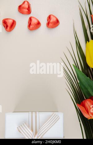 vue de dessus des bonbons au chocolat en forme de coeur enveloppés de papier d'aluminium rouge, boîte-cadeau et un bouquet de tulipes colorées sur fond blanc avec espace de copie Banque D'Images