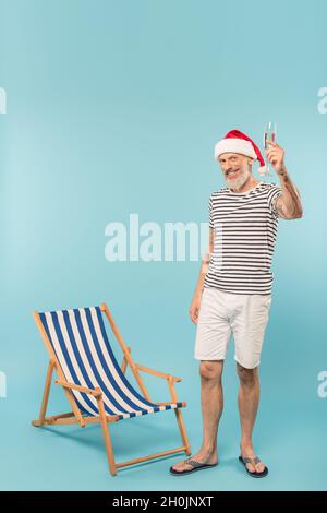 heureux homme mature dans chapeau de santa tenant une coupe de champagne près de la chaise de pont sur le bleu Banque D'Images