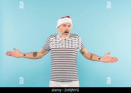 confus homme d'âge moyen dans le t-shirt stipped et chapeau de santa gesturant isolé sur bleu Banque D'Images