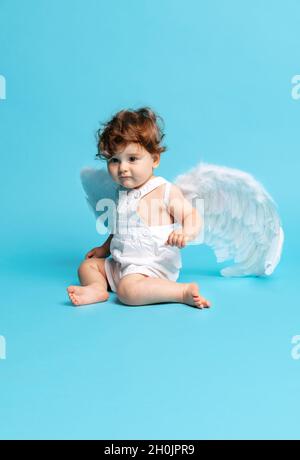 Portrait de tout-petit garçon mignon, bébé en barboteuse blanche avec ailes d'ange isolées sur fond bleu studio. Banque D'Images