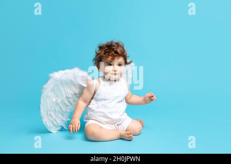 Portrait de tout-petit garçon mignon, bébé en barboteuse blanche avec ailes d'ange isolées sur fond bleu studio. Banque D'Images