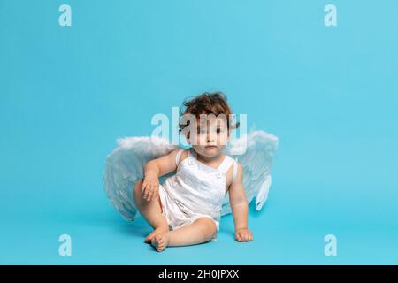 Portrait de tout-petit garçon mignon, bébé en barboteuse blanche avec ailes d'ange isolées sur fond bleu studio. Banque D'Images