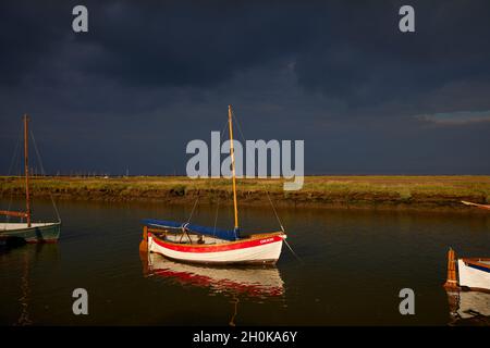 Le soleil de la fin de l'après-midi illumine un petit bateau à voile nommé « Orion » amarré à Morston Creek, sur le sentier de la côte nord de Norfolk, avec un ciel sombre avant une tempête Banque D'Images