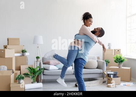 Aimant mari noir du millénaire soulevant femme excitée, célébrant la journée mouvante, amusez-vous dans la chambre moderne Banque D'Images