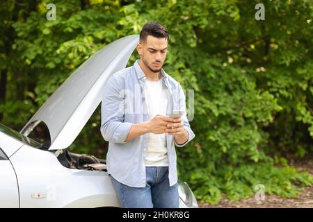 Un chauffeur de type moyen-oriental perplexe debout près d'une voiture brisée, en utilisant un téléphone cellulaire Banque D'Images