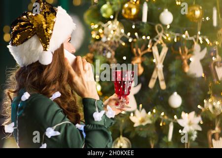 Noël.Vue de derrière une femme avec chapeau de père Noël et verre en robe verte en utilisant un smartphone près de l'arbre de Noël dans la maison. Banque D'Images