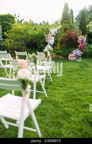 Cérémonie de mariage dans le magnifique jardin paysagé entre pelouse avec arc de mariage élégant décoré de fleurs fraîches et chaises blanches à côté. Banque D'Images