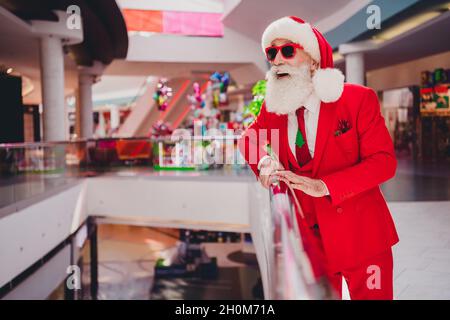 Photo du drôle impressionné santa claus porter des lunettes sombres souriant bouche ouverte regardant loin à l'intérieur du centre commercial Banque D'Images