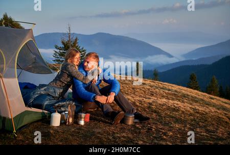 Jeunes couples amoureux randonneurs explorant de nouveaux endroits ensemble.Belle femme et homme passant du temps sur la nature dans le camping de montagne assis près de la tente touristique sur la colline herbeuse. Banque D'Images