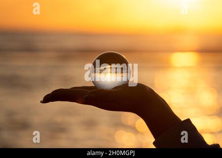 Magnifique paysage marin reflété sur une boule de cristal dans la main d'une femelle au coucher du soleil Banque D'Images