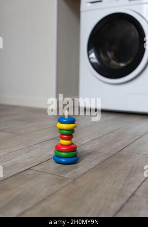 Une pyramide multicolore mal assemblée pour enfants se trouve sur le sol de la buanderie, contre le fond d'une machine à laver Banque D'Images