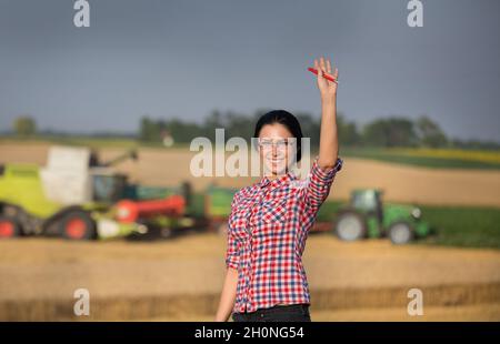 Jolie jeune femme debout sur un champ de blé pendant la récolte avec la moissonneuse-batteuse déchargeant les récoltes dans la remorque du tracteur Banque D'Images