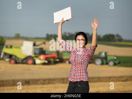 Jolie jeune femme debout sur un champ de blé pendant la récolte avec la moissonneuse-batteuse déchargeant les récoltes dans la remorque du tracteur Banque D'Images