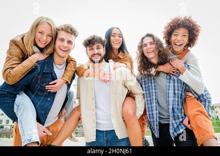 Jeunes amis ayant du plaisir à l'extérieur - six étudiants à l'extérieur, des hommes portant deux filles sur leur dos Banque D'Images