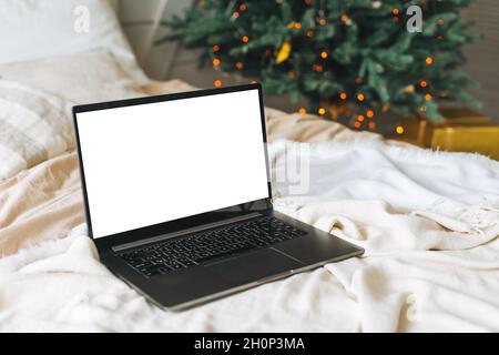 Ordinateur portable ouvert avec écran blanc sur le lit dans la chambre avec arbre de Noël décoré à la maison, maquette Banque D'Images