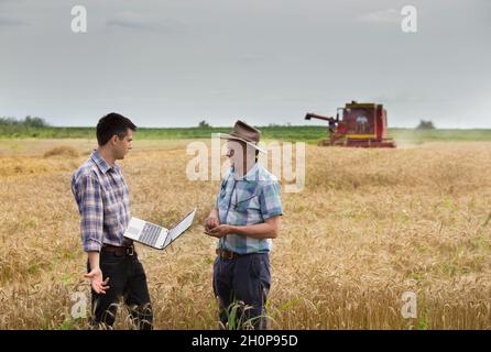 Deux agriculteurs se tenant sur un champ de blé lors de la récolte avec une moissonneuse-batteuse travaillant en arrière-plan Banque D'Images