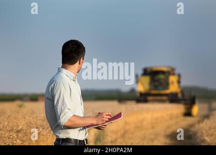 Vue arrière d'un jeune agriculteur avec ordinateur portable debout sur un champ de blé pendant que la moissonneuse-batteuse fonctionne en arrière-plan Banque D'Images