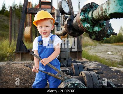 Adorable petit garçon regardant la caméra et souriant tout en serrant le boulon sur le tuyau dans le champ de pétrole.Mignon enfant dans la construction casque à l'aide d'une clé industrielle tout en réparant le tuyau de cric de pompe à l'extérieur. Banque D'Images