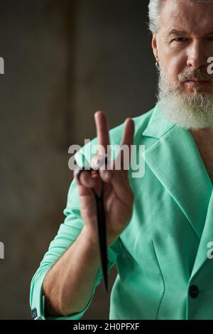 Gros plan d'un homme d'âge moyen élégant avec une barbe vêtue d'une tenue colorée, regardant l'appareil photo, montrant le signe de paix tout en tenant des ciseaux de barbier pointus Banque D'Images