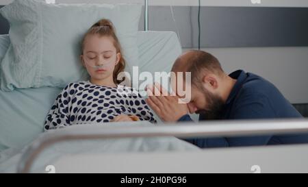 Un père inquiet priant pour une fille malade santé de rétablissement après avoir subi une chirurgie médicale dans la salle d'hôpital. Petit enfant avec tube nasal à oxygène dormant pendant l'examen de la maladie Banque D'Images